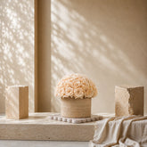 A dome arrangement of premium champagne preserved roses in a textured cylindrical travertine vase, styled on a stone pedestal with soft warm shadows for luxury home decor.